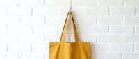 Yellow canvas tote bag hanging on white brick wall, eco-friendly reusable shopping bag mockup for branding and design projects.