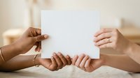 Diverse hands holding blank white paper together on wooden table, teamwork and collaboration concept for business presentations and corporate messaging.