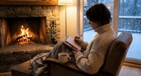 Woman reading book by fireplace in cozy living room with warm lighting and winter view outside window. Relaxing evening at home concept.