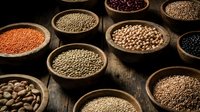 Assorted legumes and grains in wooden bowls on dark surface, including lentils, chickpeas, beans and seeds for cooking and nutrition.