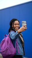 Happy young African American woman student with braids taking selfie with smartphone wearing denim jacket and purple backpack against blue wall.