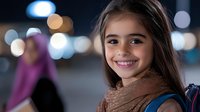 Young student with bright smile wearing brown scarf and backpack against blurred city lights background, expressing joy and confidence in evening urban setting.
