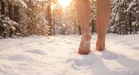 Barefoot woman walking through fresh snow in winter forest with warm sunlight filtering through trees creating magical atmosphere for wellness concepts.