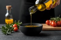 Pouring golden olive oil from bottle into black ceramic bowl with spices on dark background, fresh tomatoes and herbs for Mediterranean cuisine preparation.