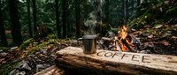 Steaming metal mug on a rustic log engraved with coffee, beside a glowing campfire in a misty forest setting.