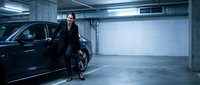 Professional businesswoman in black suit standing beside luxury car in modern underground parking garage with concrete walls and fluorescent lighting.