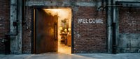 Rustic brick warehouse exterior with open metal door glowing warmly inside, welcome sign on aged wall, industrial urban entrance concept.