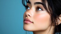 Young Asian woman with natural makeup and glowing skin looking up against blue background, dark hair styled with loose strands framing face. Professional beauty portrait.