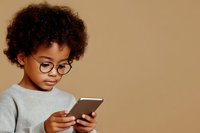 Young African American child wearing glasses and gray sweater focused on digital tablet against brown background, modern education concept.