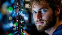 Young bearded Caucasian man with intense blue eyes in dramatic lighting against dark background with colorful bokeh lights, conveying confidence and determination.