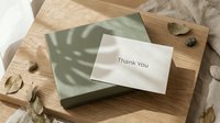 Elegant thank you card resting on a sage green gift box with natural leaf shadows on a wooden surface, surrounded by dried leaves.