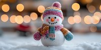 Cheerful knitted snowman toy with colorful winter hat and scarf on snow against blurred warm bokeh lights background, festive holiday decoration.