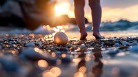 Bare feet walking on pebble beach at golden sunset with seashell in foreground, creating magical bokeh effect on wet stones near ocean shore.