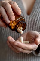Close-up of hands pouring white capsules from amber glass bottle into palm, wearing gray knit sweater. Medical supplement concept for healthcare marketing.