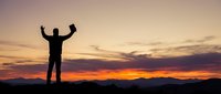 Silhouette of successful businessman celebrating victory with raised arms against dramatic sunset sky over mountain landscape for motivation concepts.