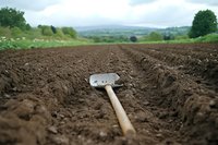 Garden hoe lying on freshly plowed soil rows in rural countryside with rolling hills and green vegetation in background, agricultural farming concept.