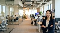 Young woman of South Asian descent wearing professional black blazer and beige blouse, standing confidently with crossed arms in modern open-plan office environment with colleagues working at desks.