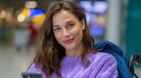 Young woman with brown wavy hair wearing purple sweater and blue jacket smiles warmly at camera in indoor setting with soft bokeh background lighting.