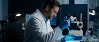 Male scientist in white lab coat and blue gloves conducting research with microscope and test tubes in modern laboratory setting. Suitable for healthcare, medical research, and science education.