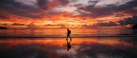 Silhouette of male jogger running along sandy beach at dramatic golden sunset with vibrant orange and red sky reflected in wet sand, ideal for fitness and wellness content.