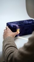 Person opening a luxurious deep blue velvet jewelry box with gold hinges, hands wearing a silver ring, elegant gift concept.