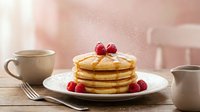 Fluffy golden pancakes topped with fresh raspberries and powdered sugar on white plate with coffee cup and milk pitcher on wooden table in cozy kitchen.