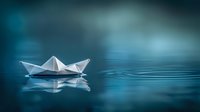 White paper boat floating on calm blue water surface with reflection and soft bokeh background, creating peaceful minimalist scene for concepts of journey and hope.