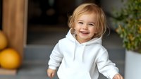 Cheerful toddler girl with blonde hair wearing white hoodie smiles brightly while playing at home, natural indoor lighting creates warm atmosphere.