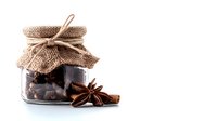 Glass jar filled with dark chocolate pieces, decorated with rustic burlap top and string, star anise and cinnamon sticks on white background, isolated food composition.
