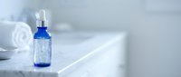 Blue glass dropper bottle with clear serum on white marble surface in minimalist bathroom setting for skincare and beauty product applications.