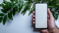 Mockup of smartphone with blank white screen held in male hand against white background with green leaves and branches, copy space for app design.