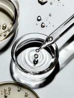 Clear liquid droplet falling from metal dropper into glass container creating ripple effect on white surface with scattered drops and petri dishes in view.
