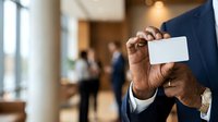 Professional man with dark skin tone holding blank white business card in modern office lobby with colleagues in background. Corporate networking and business identity concept.