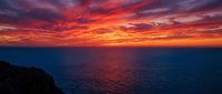 Dramatic ocean sunset with vibrant red and orange sky reflecting on calm water, silhouetted coastal cliffs in foreground, ideal for travel and nature backgrounds.