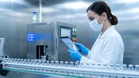 Female scientist in white lab coat and mask operating automated laboratory equipment for pharmaceutical research and quality control testing.