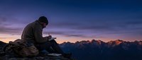 Male hiker using smartphone on mountain summit during golden hour sunrise with dramatic alpine peaks and purple sky for outdoor adventure content.