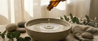 Honey dripping from wooden dipper into ceramic bowl creating ripples, surrounded by eucalyptus leaves and smooth stones in warm natural sunlight.