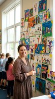 Smiling Caucasian female teacher stands proudly beside a colorful wall of childrens artwork in a bright elementary school classroom.