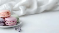 Delicate French macarons in soft pastel pink and lavender shades arranged on white ceramic plate with fresh lavender sprig, against light fabric background.