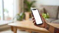 Young adult with dark skin tone holding smartphone with blank white screen in modern living room with natural light, suitable for app mockups and mobile technology marketing.