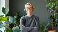Young Caucasian businesswoman wearing glasses and grey sweater stands confidently with arms crossed near green plants in modern office space, expressing professional attitude.