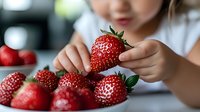 Young Asian child holding fresh ripe strawberry, close up view of hands with red berries on white table, selective focus on fruit, healthy eating lifestyle concept.