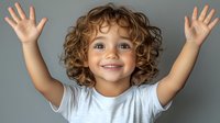 Happy mixed race child with curly hair raising hands up in joyful expression against gray background, wearing white t-shirt, showing excitement and positive emotions.