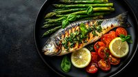 Grilled sea bass with fresh herbs served with asparagus, cherry tomatoes, and lemon slices on black plate. Healthy Mediterranean cuisine.