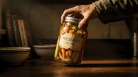 Male hand holding glass jar with preserved vegetables and pickled food in warm rustic kitchen setting with books and bowls for home canning and food storage.