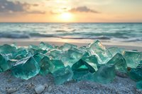 Turquoise sea glass pieces scattered on sandy beach at sunset, waves and golden sky in background, creating magical coastal atmosphere.