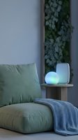 Cozy modern living room corner with green armchair, glowing LED orb lamp and wireless speaker on wooden side table, blue throw blanket, plant wall.