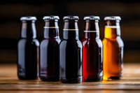 Assorted craft beer bottles showing different varieties from dark stout to light lager, arranged in row on wooden surface with condensation droplets.