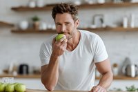 Handsome Caucasian man in white t-shirt eating green apple in modern kitchen with wooden shelves, promoting healthy lifestyle and nutrition.