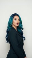 Professional young woman with vibrant teal blue hair and dark lipstick wearing black blazer against neutral background for business portrait.